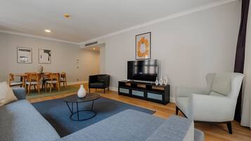 Ein modernes Wohnzimmer mit einer Couch, einem Couchtisch, einem Fernseher auf einem schwarzen Standfuß, einem Essbereich mit Stühlen und einem Tisch sowie einem hellen Sessel.