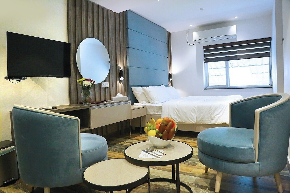Hotellihuone, jossa on sänky, televisio, työpöytä, kaksi tuolia, pyöreä pöytä ja kulho täynnä hedelmiä.