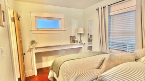 A bedroom with a wooden floor, a bed with striped bedding, a desk with a mirror, and a window with blinds. - Coastal Beach Bungalow walking distance to beach, coffee, and restaurants (Redondo Beach)