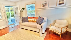 A living room with a white sofa, a small round table, and a wooden chair. There are two windows with blinds, a door leading outside, and a rug on the floor. - Coastal Beach Bungalow walking distance to beach, coffee, and restaurants (Redondo Beach)