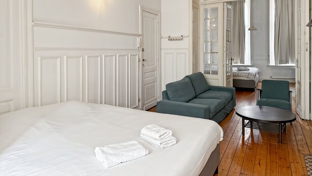 A hotel room with a large bed, a green sofa, a small table, and a view through a large window. - Prime Location Apartment Châtelain Saint-gilles Quiet & Central (Brussels)