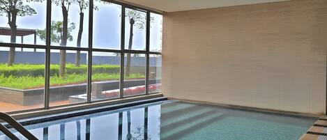 An indoor swimming pool with clear water, a wooden deck, and a view of greenery and a building through large windows.