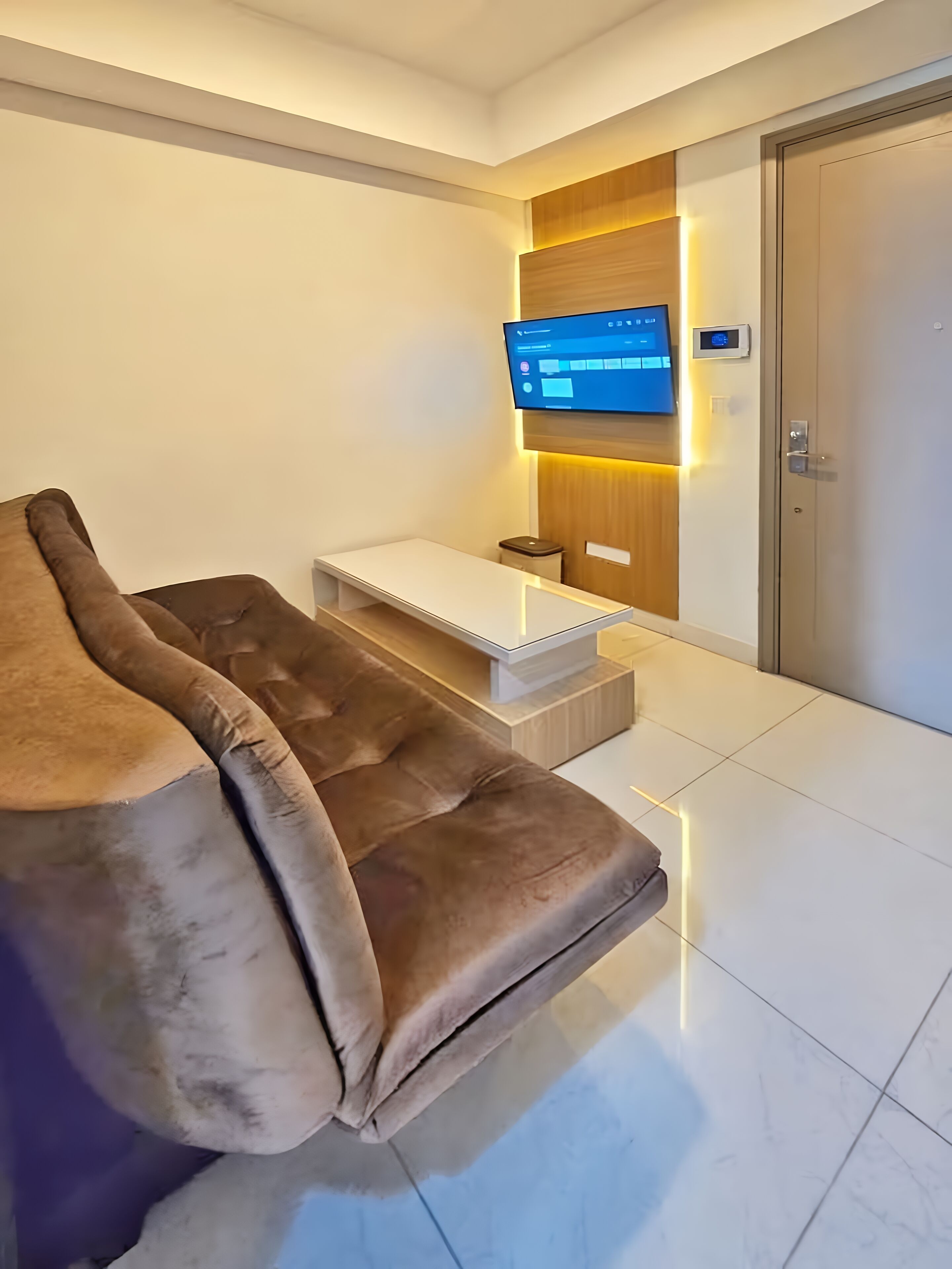 A modern hotel room with a brown leather sofa, a flat-screen TV mounted on the wall, a small table, and a door with a digital lock.