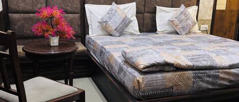 Une chambre avec une tête de lit en bois, un lit recouvert d’une literie à motifs, une chaise, une petite table sur laquelle est posé un vase de fleurs, et un tapis sur le sol.