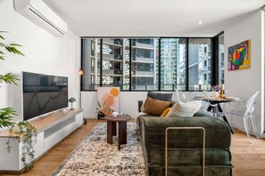 Ein modernes Wohnzimmer mit großem Fernseher, einer Couch, einem Esstisch und Blick auf hohe Gebäude.