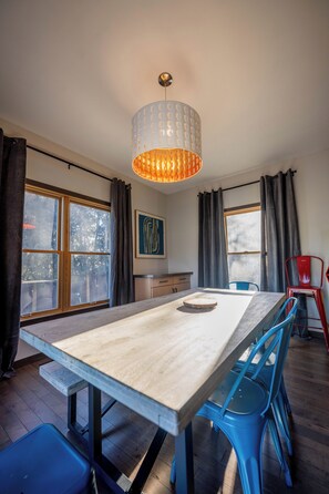 Un espace repas avec une longue table, des chaises bleues, un luminaire suspendu et de grandes fenêtres avec des rideaux.