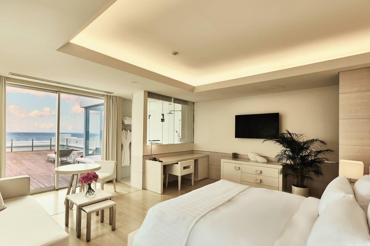 A modern hotel room with a large bed, a desk, a television, and a view of the ocean.