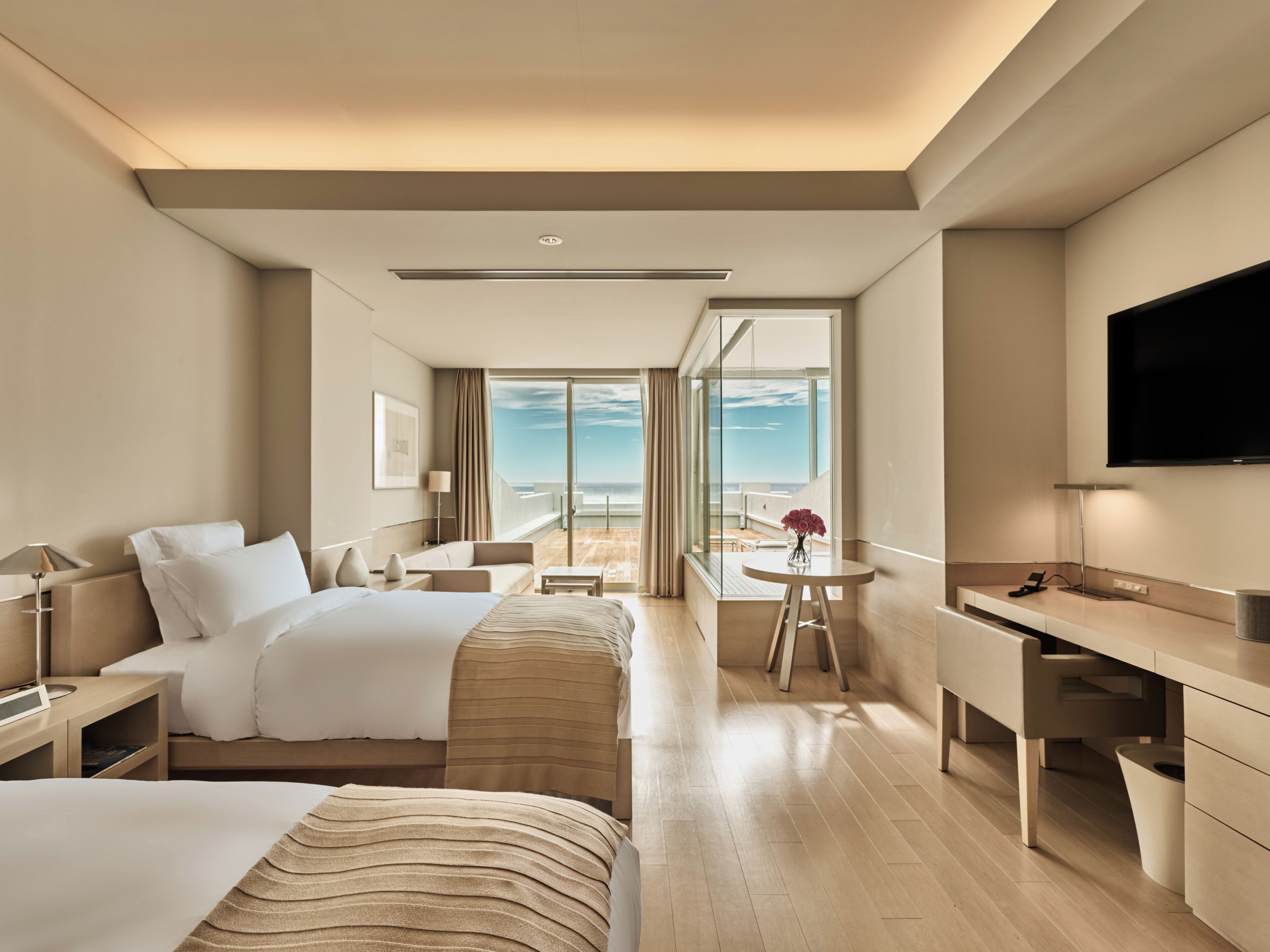 A hotel room with a bed, a desk, a chair, a TV, and a balcony with a view of the ocean.