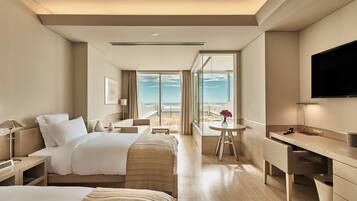 A hotel room with a bed, a desk, a chair, a TV, and a balcony with a view of the ocean.