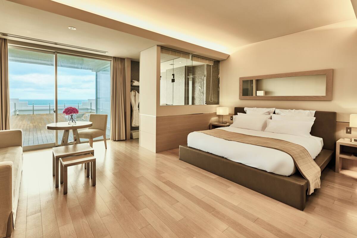 A modern hotel room with a large bed, a seating area with a table, a bathroom with a shower, and a balcony with a view.