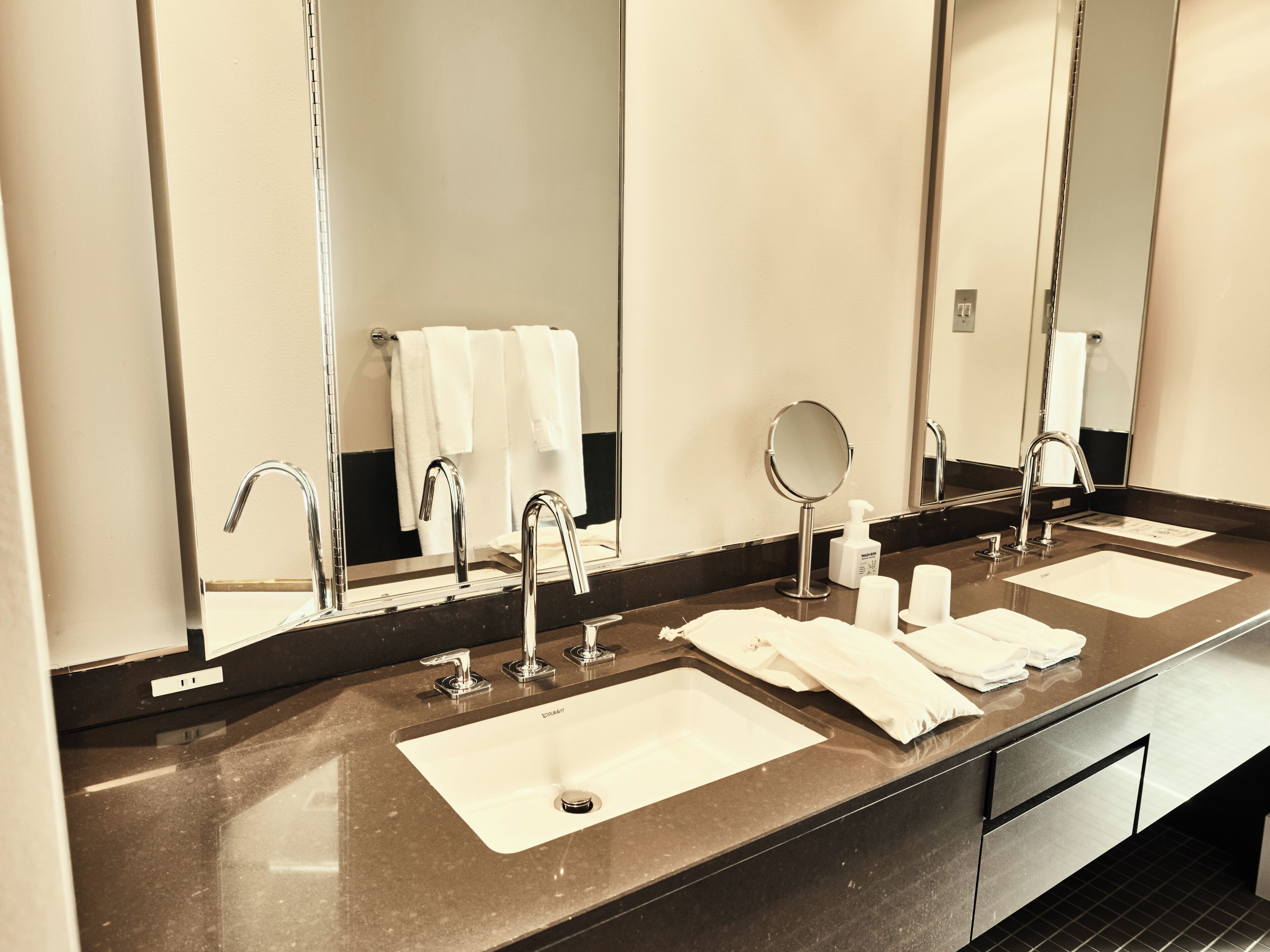 A bathroom with two sinks, a mirror, a towel rack, and a soap dispenser.