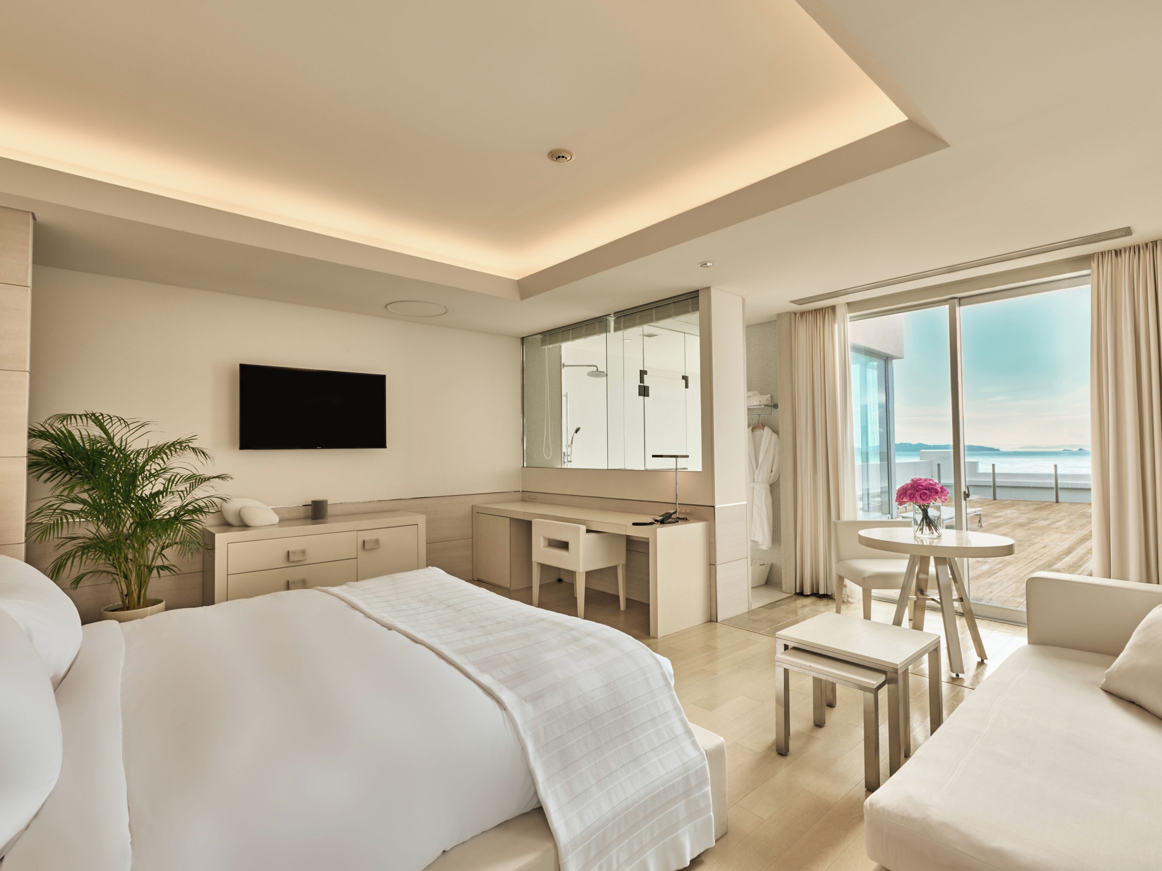 A modern hotel room with a bed, a desk, a chair, a small table, a plant, a TV, and a large window with a view.