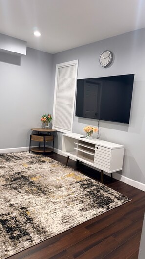 A modern living room with a large flat-screen TV mounted on the wall, a white console table with drawers, a round wooden table with a vase of flowers, and a patterned rug on the floor. - Clean & Furnished suite near major hospitals. Move-In Ready for Professionals. (Newark)