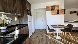 A modern kitchen with dark countertops, a white refrigerator, and a potted orchid. There is a dining area with a wooden table and two black chairs. The room has wooden flooring and a white door in the background. - Rainbow Apartments in Marinha Grande (Marinha Grande)