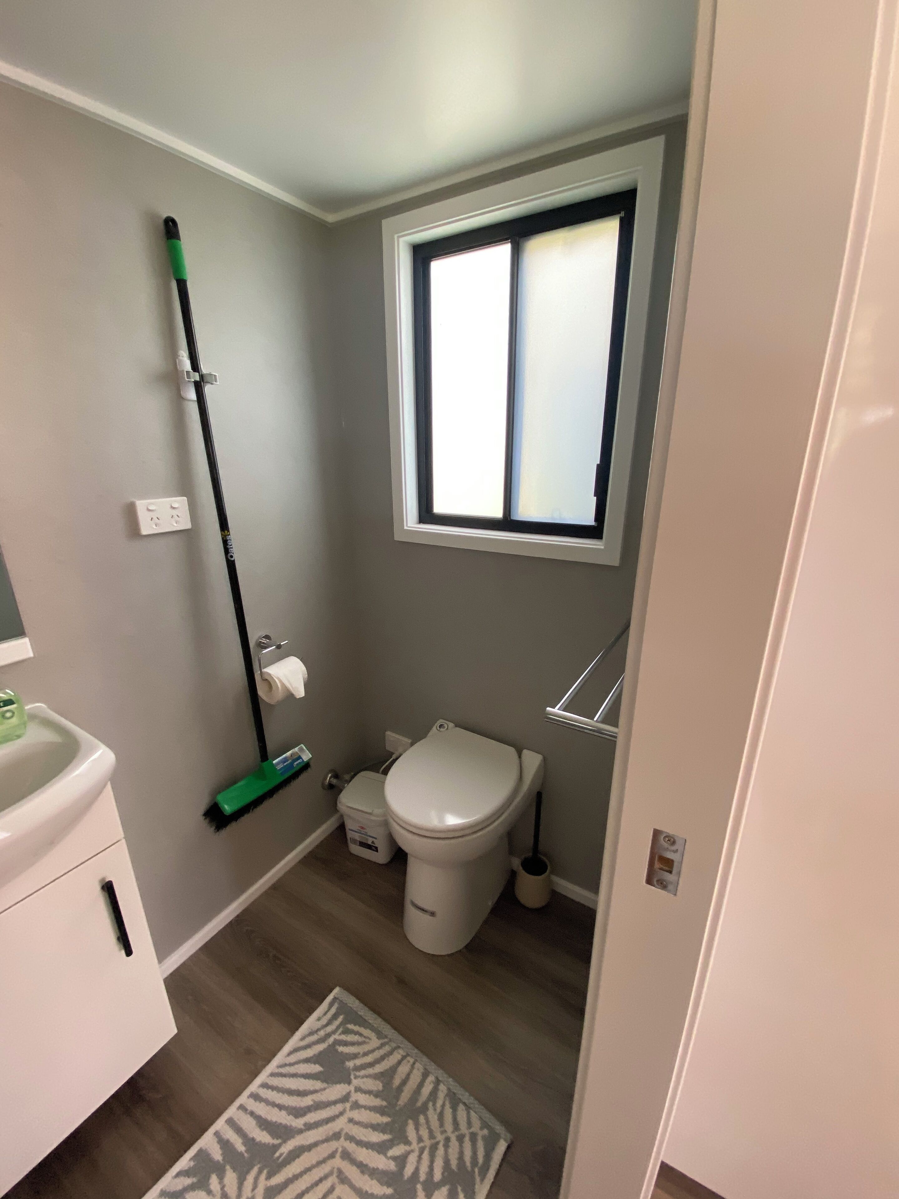 Ein kleines Badezimmer mit Toilette, Fenster, einem Mop, einem Mülleimer und einem Waschbecken.
