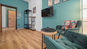 A modern living room with teal walls, a wooden floor, a green sofa with a colorful pillow, a black TV, and a small round table. - Apartment at Linden Square - Trutnov (Trutnov)