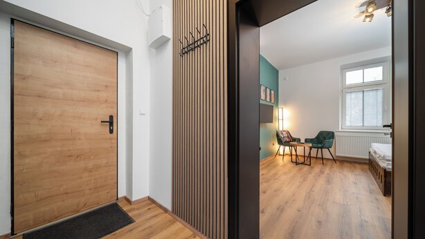 A modern room with a wooden floor, a black door, a wall-mounted coat rack, a green chair, a small table, and a bed with white bedding. - Apartment at Linden Square - Trutnov (Trutnov)