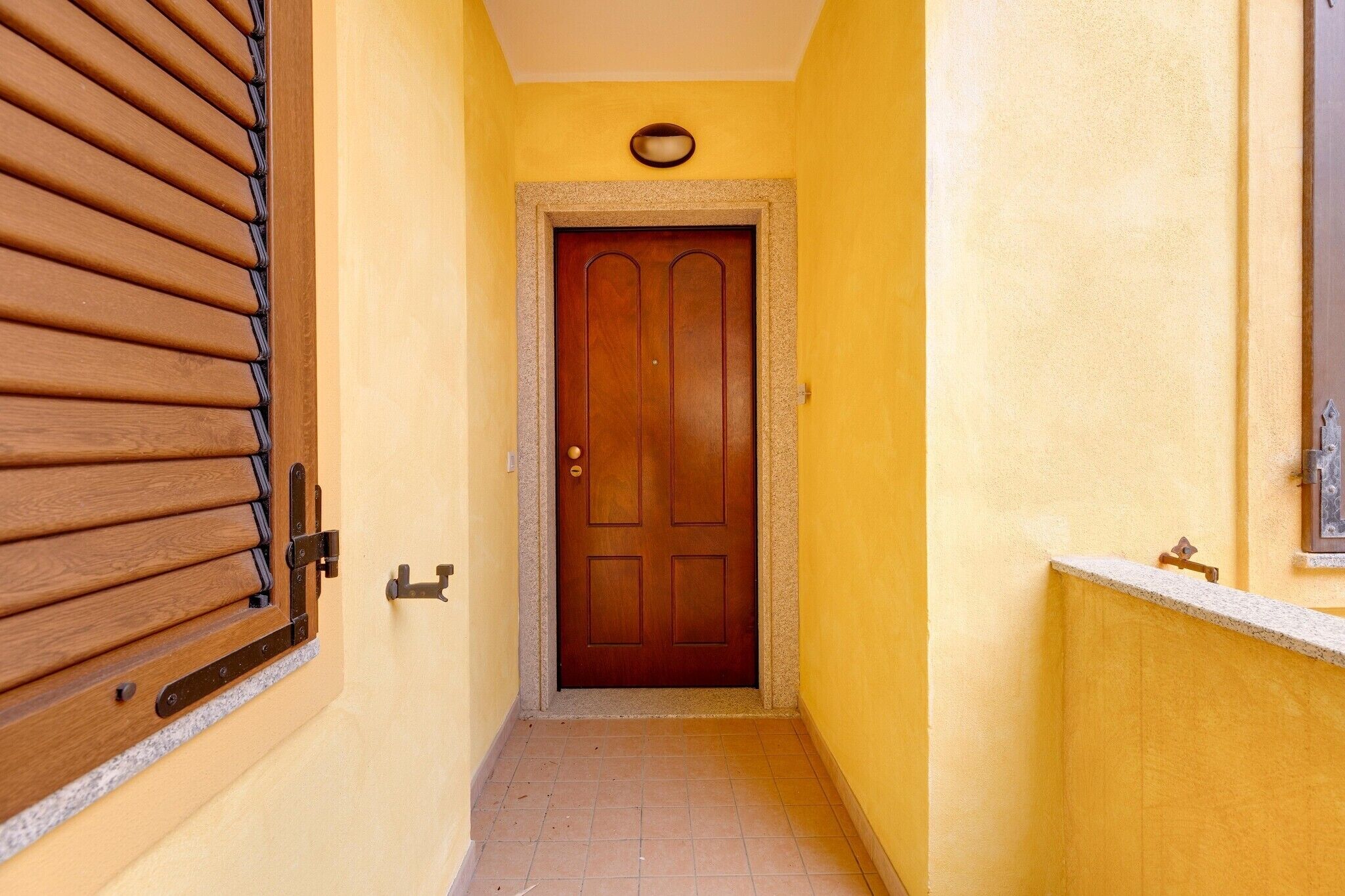 Un pasillo estrecho con una puerta de madera, una ventana con persianas y una lámpara fijada a la pared.