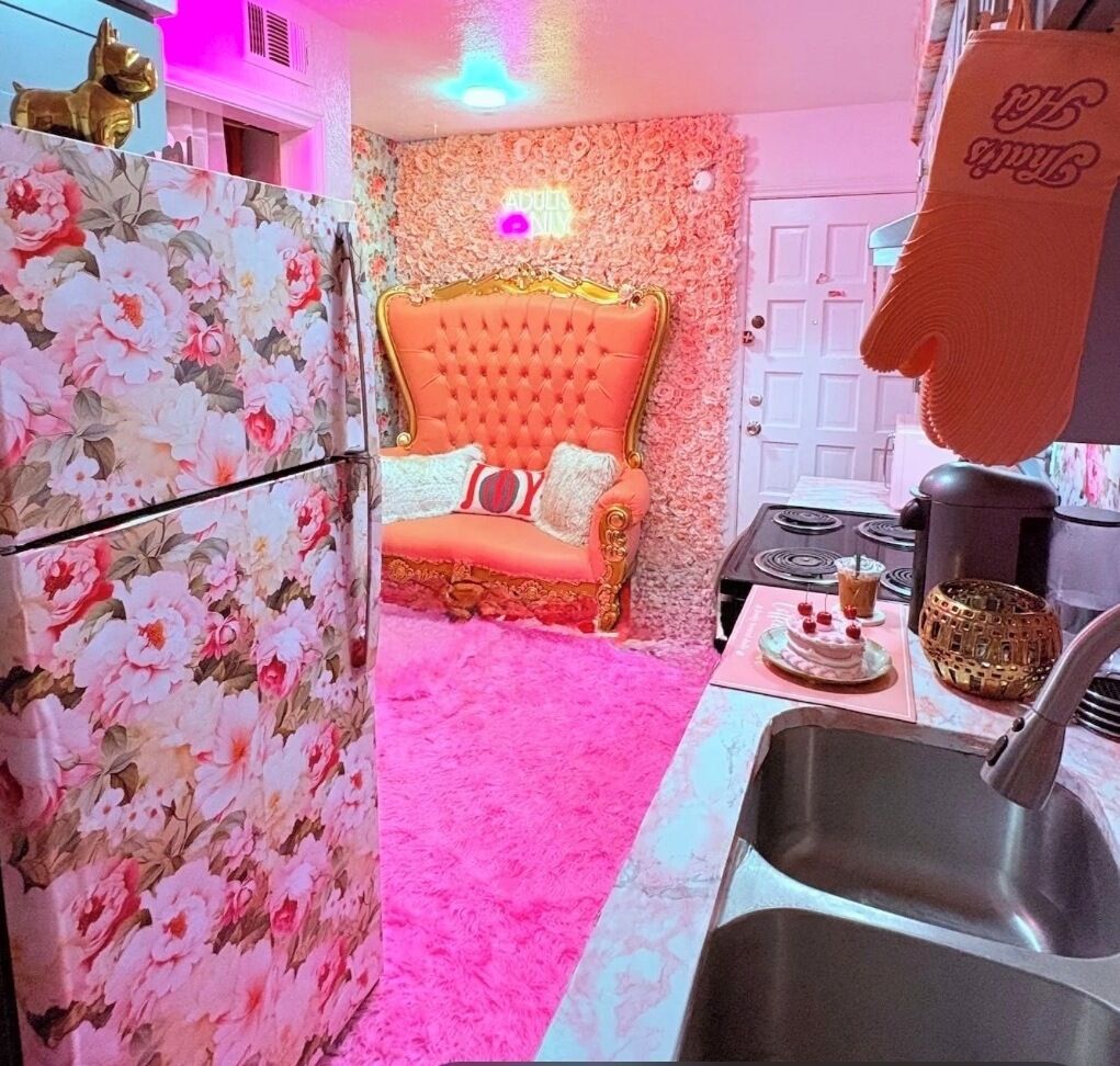 A kitchen with a pink carpet, a floral refrigerator, a golden statue, and a chair with a 'JOY' pillow.