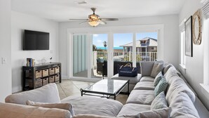 A modern living room with a sofa, coffee table, and a television. There is a ceiling fan, a sliding glass door leading to a balcony, and a wall-mounted TV. - Exclusive Beach Access + Private Pool + Sunset Views | Sandy Pelican (Seacrest)