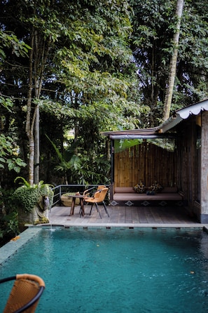 A pool with wooden deck seating, surrounded by lush greenery and trees. - Sacred Jewel by GenuineHost (Payangan)