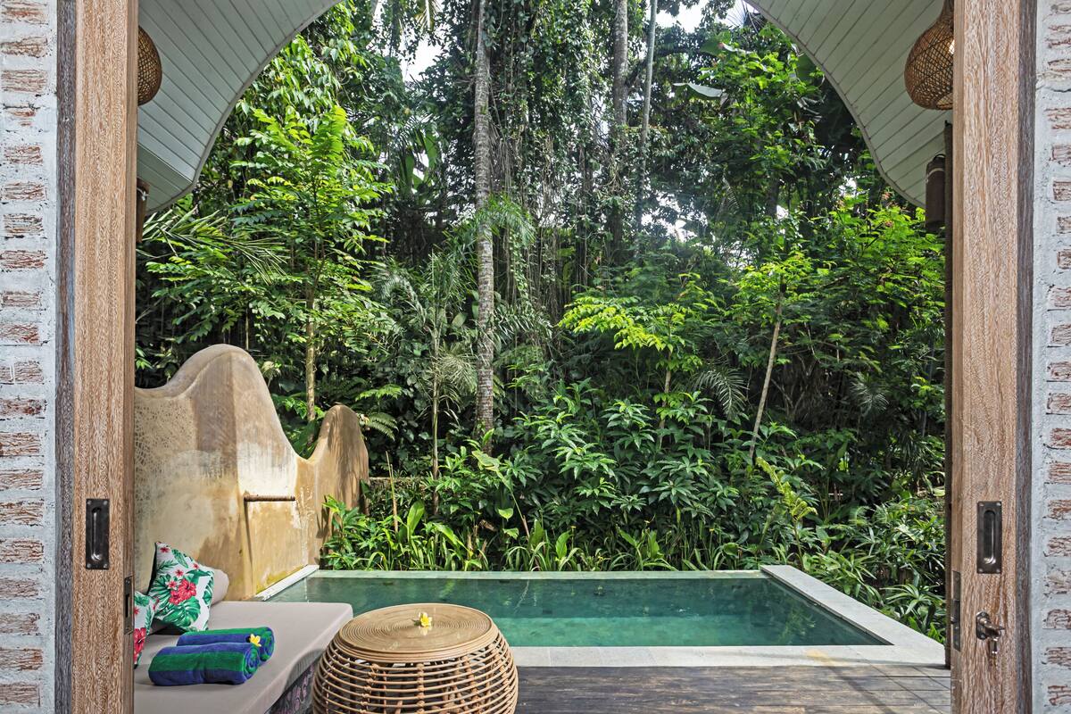 Uma área de piscina privativa com um banco de pedra, um banquinho de vime e vegetação exuberante ao fundo.