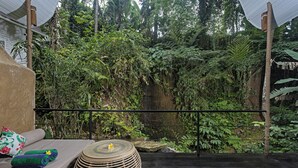 A room with a bed, a round woven stool, and a view of dense greenery. - Sacred Jewel by GenuineHost (Payangan)