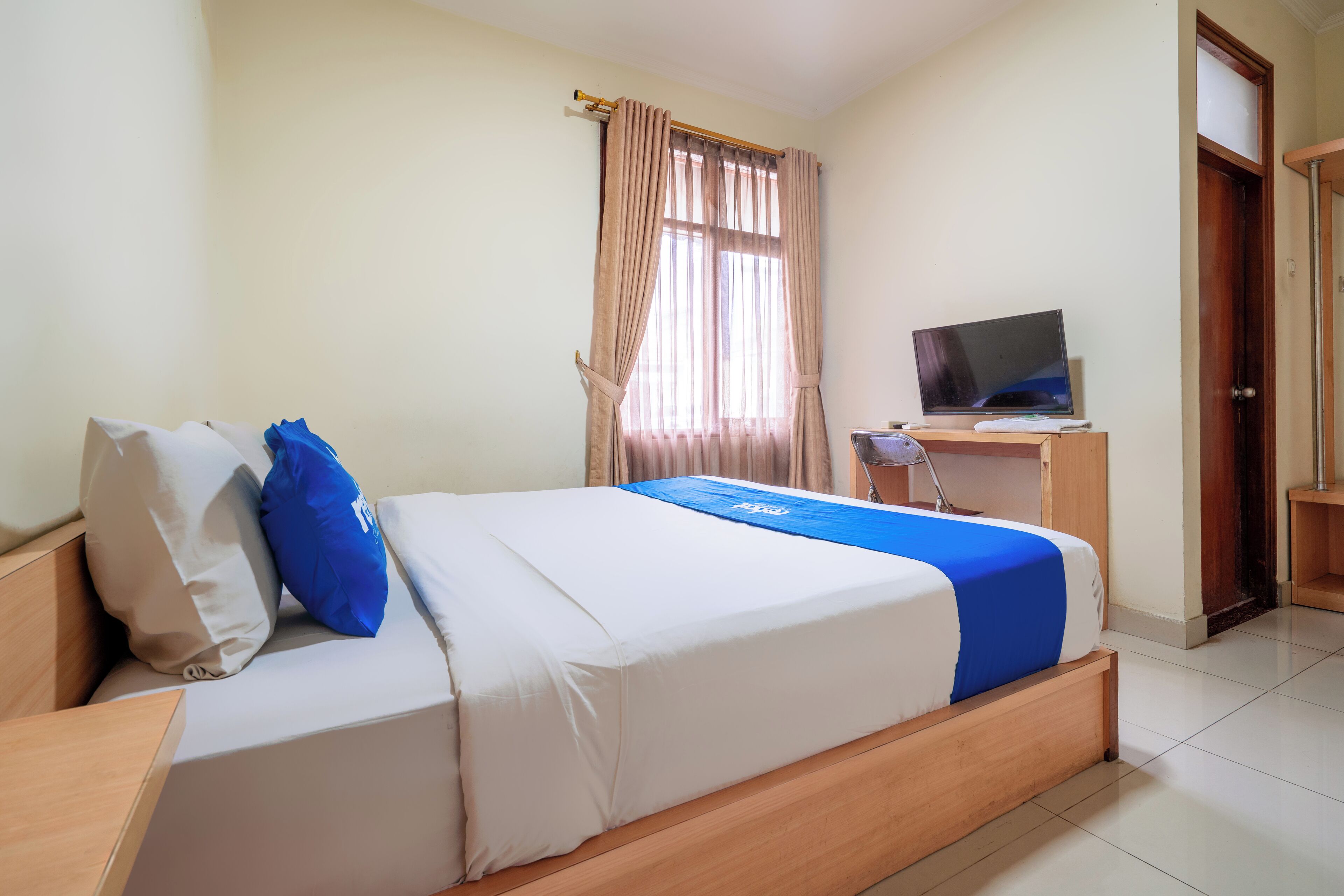 A neatly made bed with a blue and white bedspread, a television on a wooden desk, and a window with curtains.