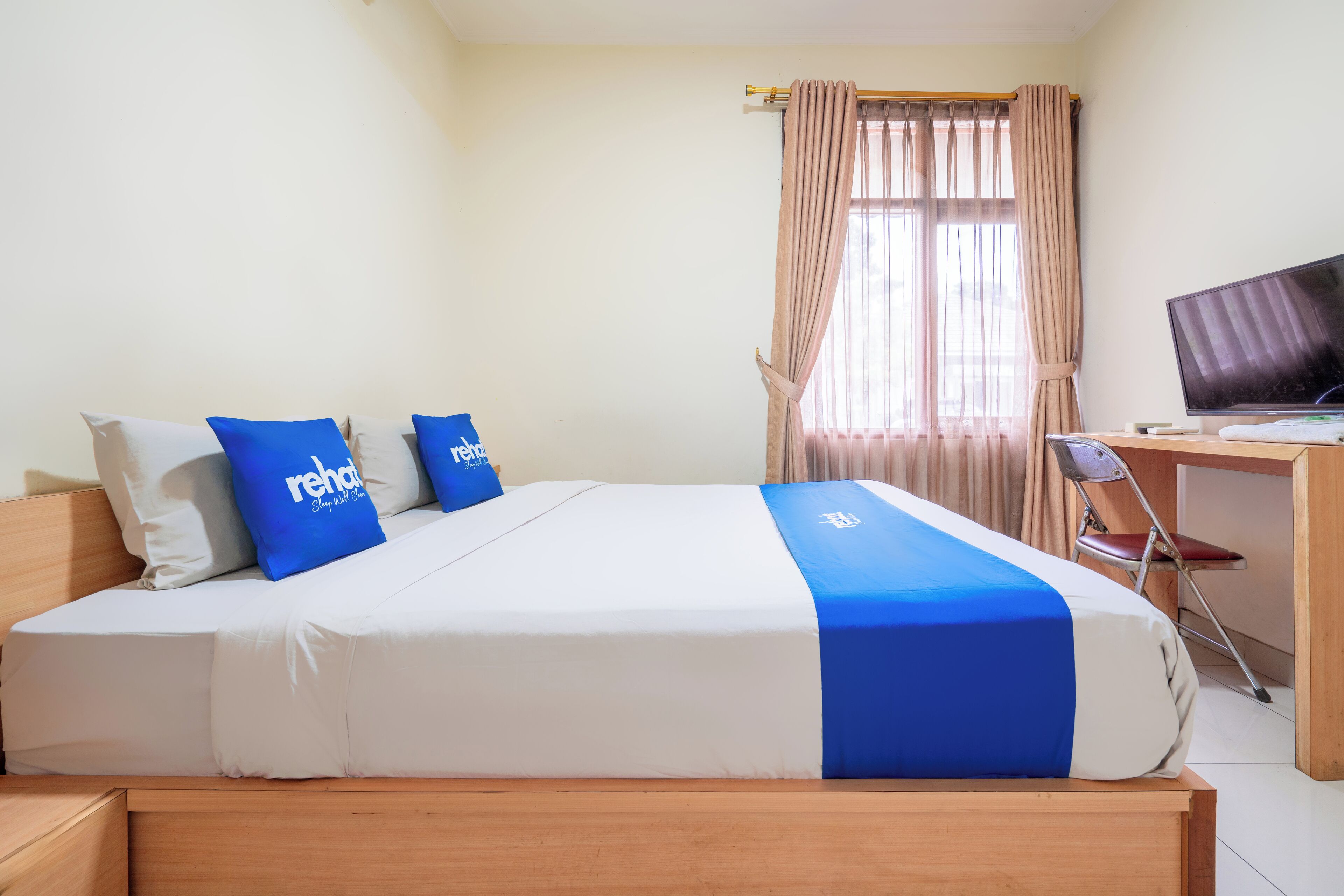 A neatly made bed with blue and white bedding, a television on a wooden desk, and a window with curtains.