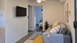 A modern living room with a sofa, a flat-screen TV mounted on the wall, a ceiling fan, and a small Christmas tree.
