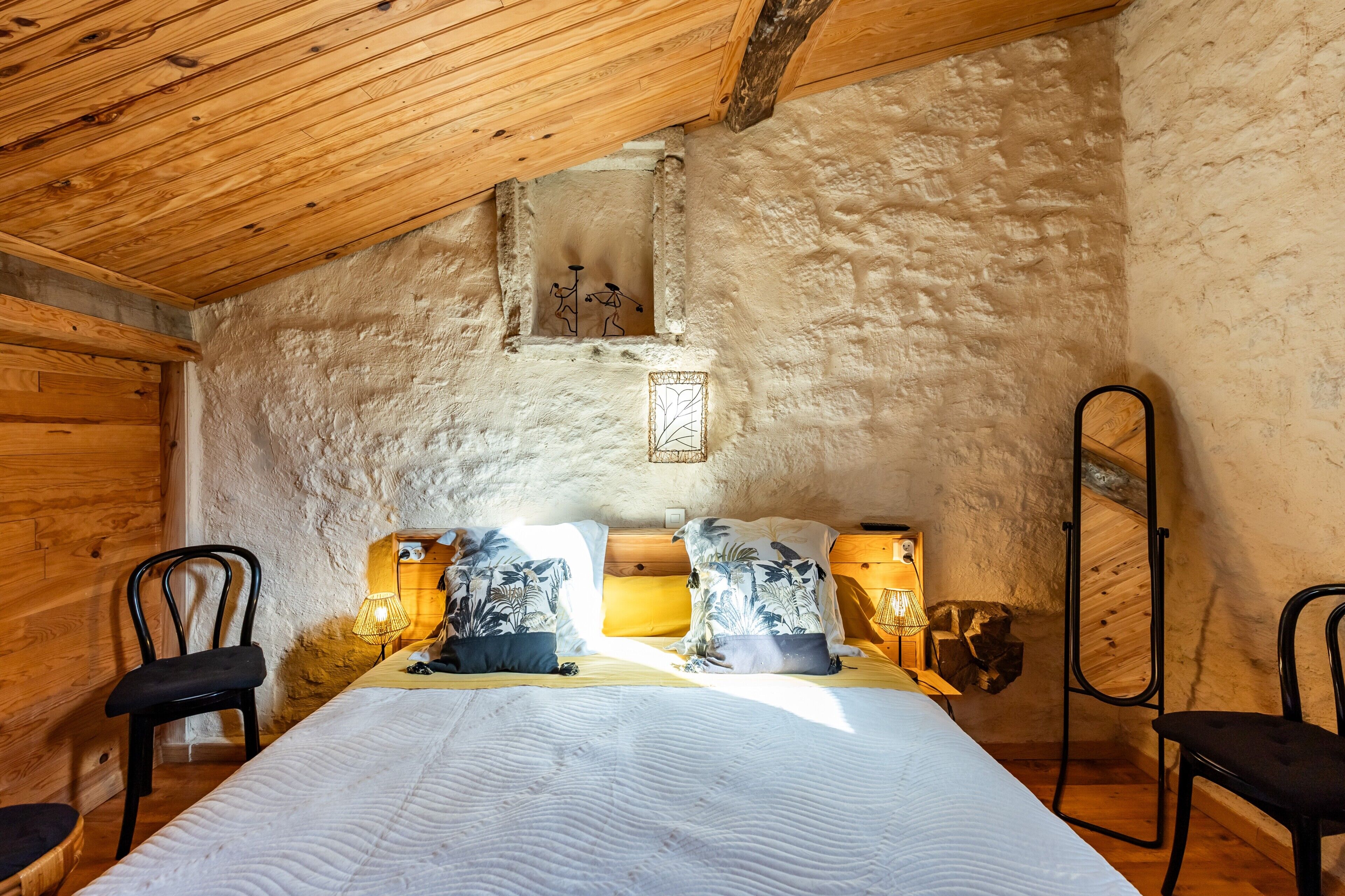 A rustic room with a wooden ceiling, a bed with patterned pillows, two chairs, a small window, and a mirror.