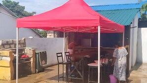 Une tente rouge est installée dans un espace extérieur doté d’un sol carrelé, entourée de chaises et de tables, et une personne se tient à proximité.
