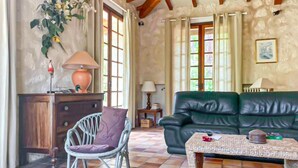 A cozy living room with a green sofa, a wooden coffee table, and a chair. There are two windows with curtains, a lamp on a side table, and a wall-mounted shelf with decorative items. - Maison accès plage direct (Les Mathes)