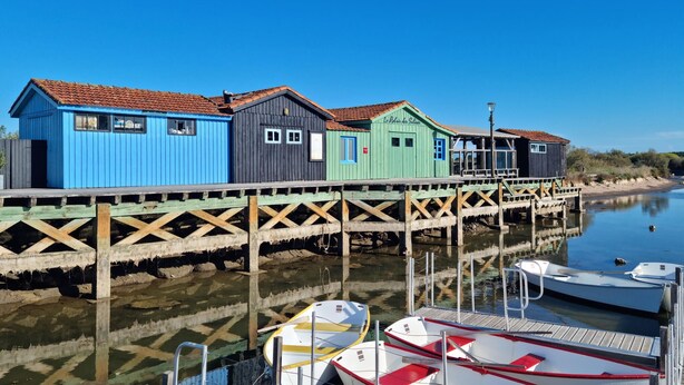 A row of colorful wooden buildings with a wooden dock extending into calm water. - Maison accès plage direct (Les Mathes)