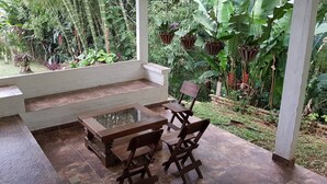 Een overdekt terras met een houten tafel en stoelen, omgeven door weelderige begroeiing en planten.