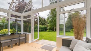 A conservatory with a glass roof, a seating area with cushions, and a view of a well-maintained garden. - Rural Norfolk Cottage welcoming 3 dogs (Hickling)