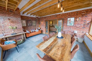 A spacious room with a large wooden dining table surrounded by chairs, a brick wall, and a cozy seating area with a sofa and cushions. - Deberri - sleeps 8 guests  in 4 bedrooms (Fressingfield)