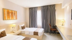 A hotel room with two beds, a desk, a chair, a lamp, a painting, and a window with curtains. - Scarlet Hotel Stasiun Bandung (Bandung)