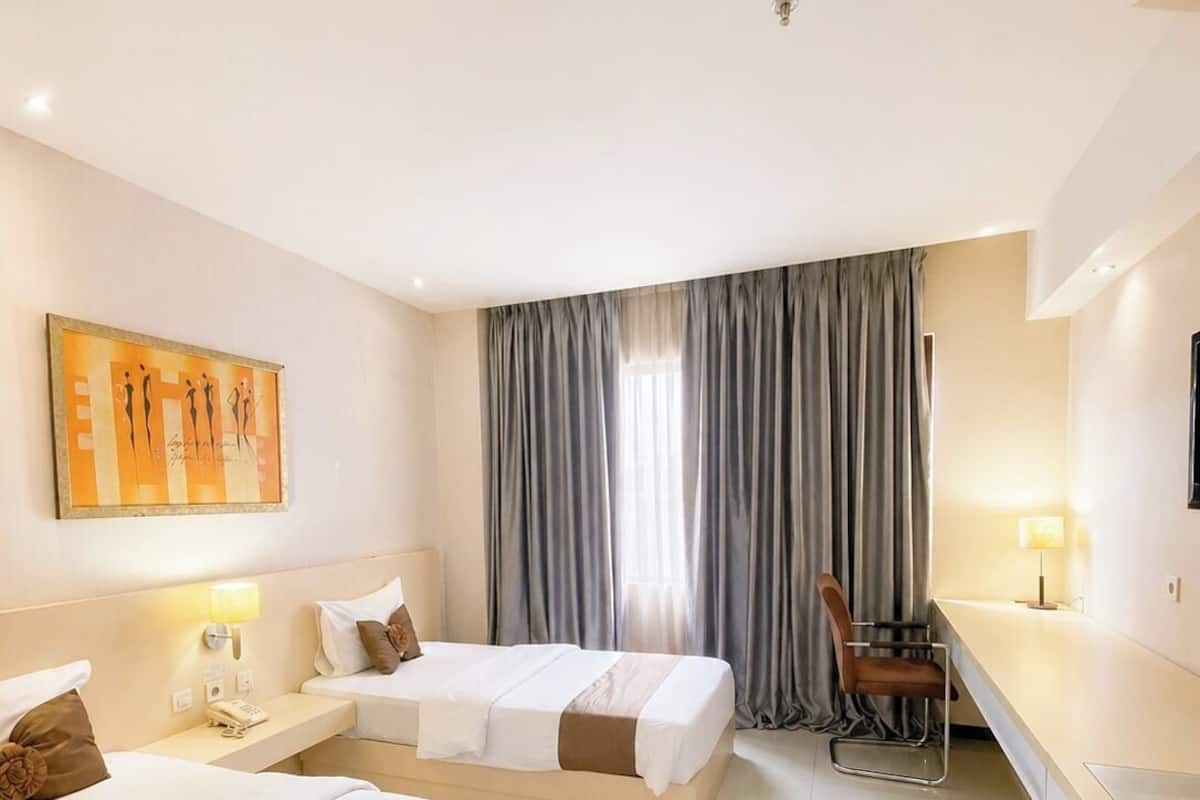 A hotel room with two beds, a desk, a chair, a lamp, a painting, and a window with curtains.