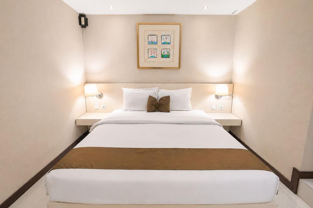 A hotel room with a large bed, two bedside lamps, a framed picture on the wall, and a wooden headboard.