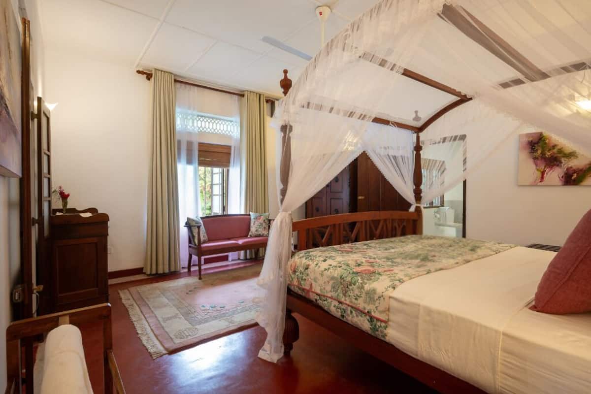 Un lit traditionnel à baldaquin, agrémenté d’une couvre-lit à motifs floraux, d’une table de chevet en bois, d’un banc et d’un miroir mural.