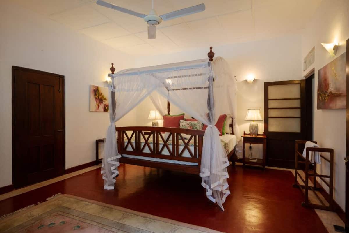 Un lit à baldaquin, un ventilateur de plafond, une porte en bois, une table de chevet avec une lampe et une étagère fixée au mur.