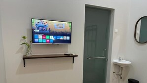 A modern room with a flat-screen TV mounted on the wall, a small shelf below it, a glass door, a sink, and a trash bin. - Cendhani Raras Gallery (Depok)