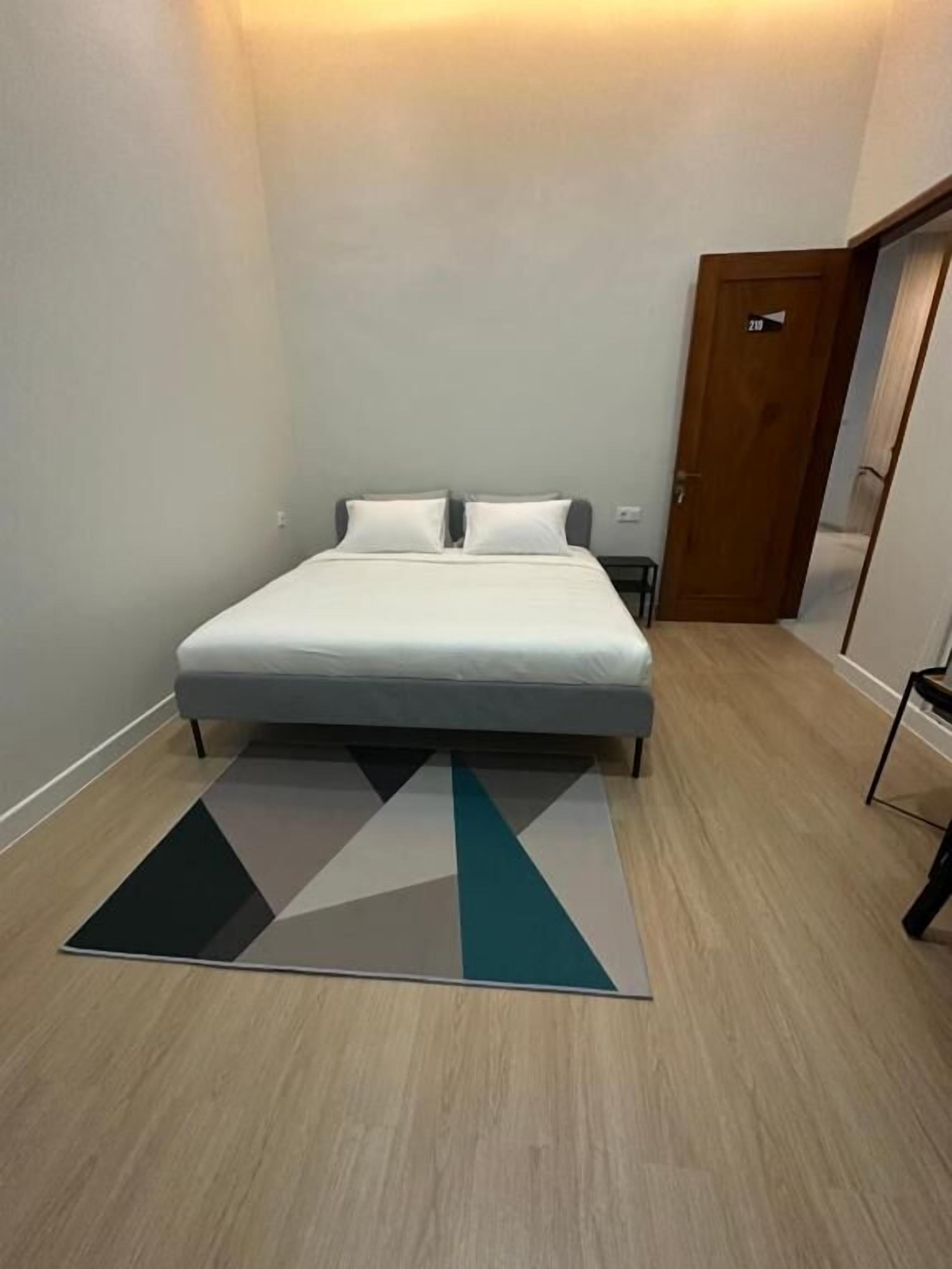 A bedroom with a bed, a geometric rug, and a door with a number.