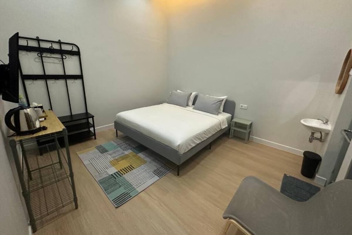 A compact hotel room with a bed, bedside tables, a small desk, a chair, a desk lamp, a wall-mounted shelf, and a bathroom with a sink and toilet.