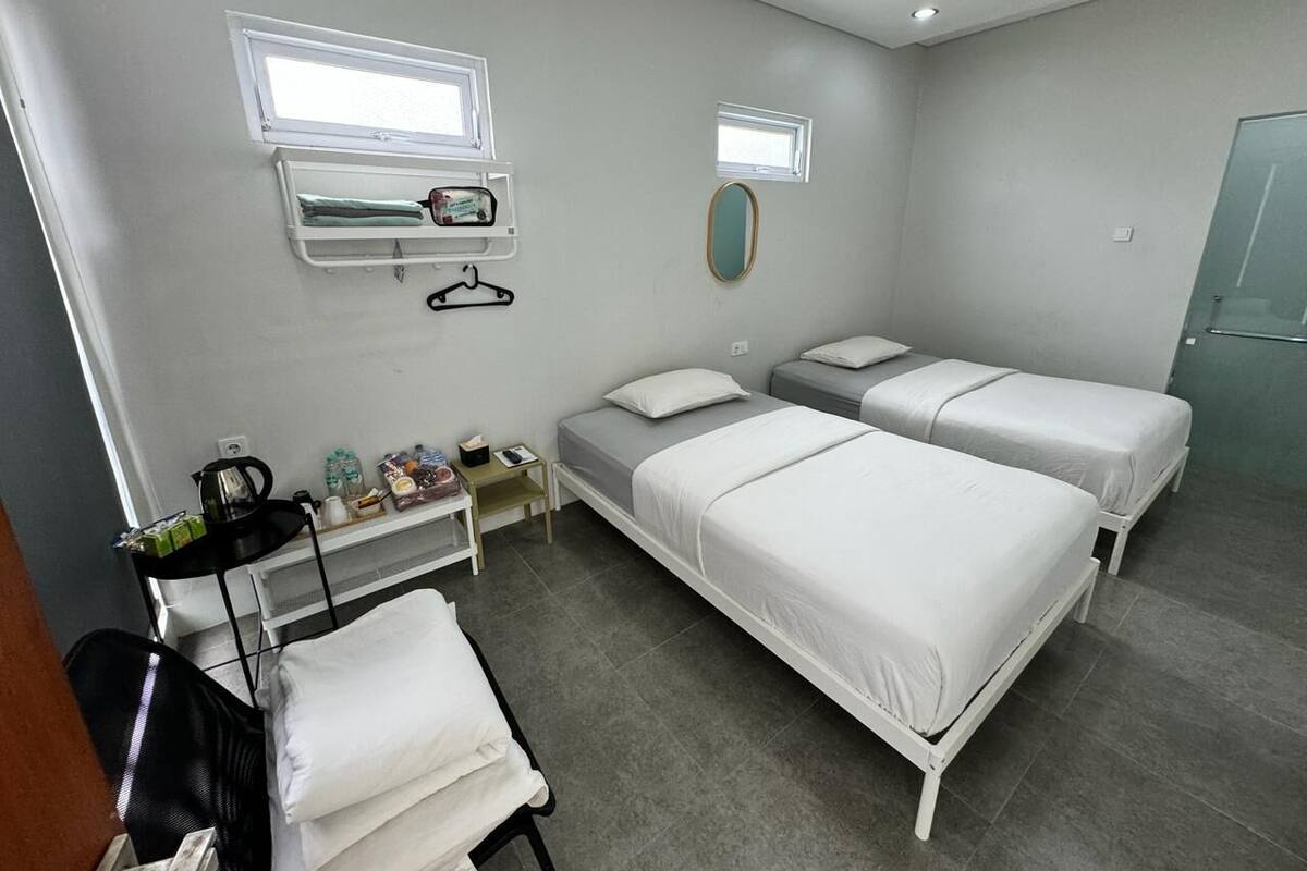 A room with two single beds, a small table with a chair, a shelf with toiletries, and a window.