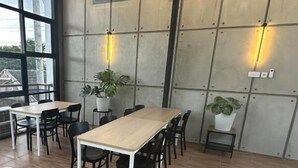 A modern dining area with wooden tables and chairs, potted plants, and a large window providing natural light. - Cendhani Raras Gallery (Depok)