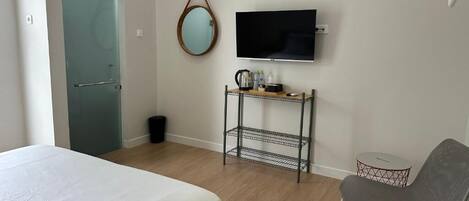 A bedroom with a bed, a TV, a small table with a kettle, a chair, and a mirror on the wall.