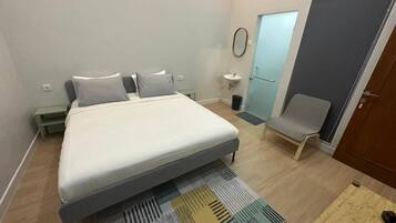 A bedroom with a bed, a chair, a small table, a sink, and a door.