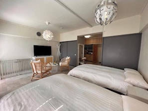 Moderni hotellihuone, jossa on kaksi sänkyä, televisio, ruokailutila tuoleineen ja pieni keittiönurkkaus.